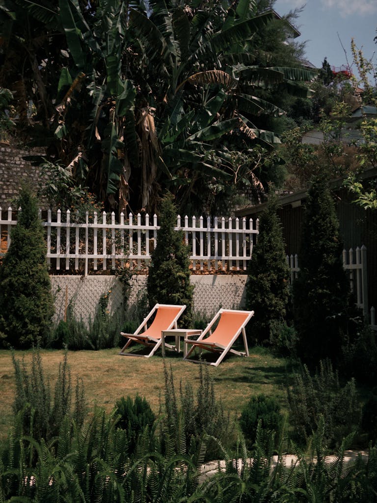 Peaceful backyard scene with two deck chairs amidst lush greenery and a white picket fence.