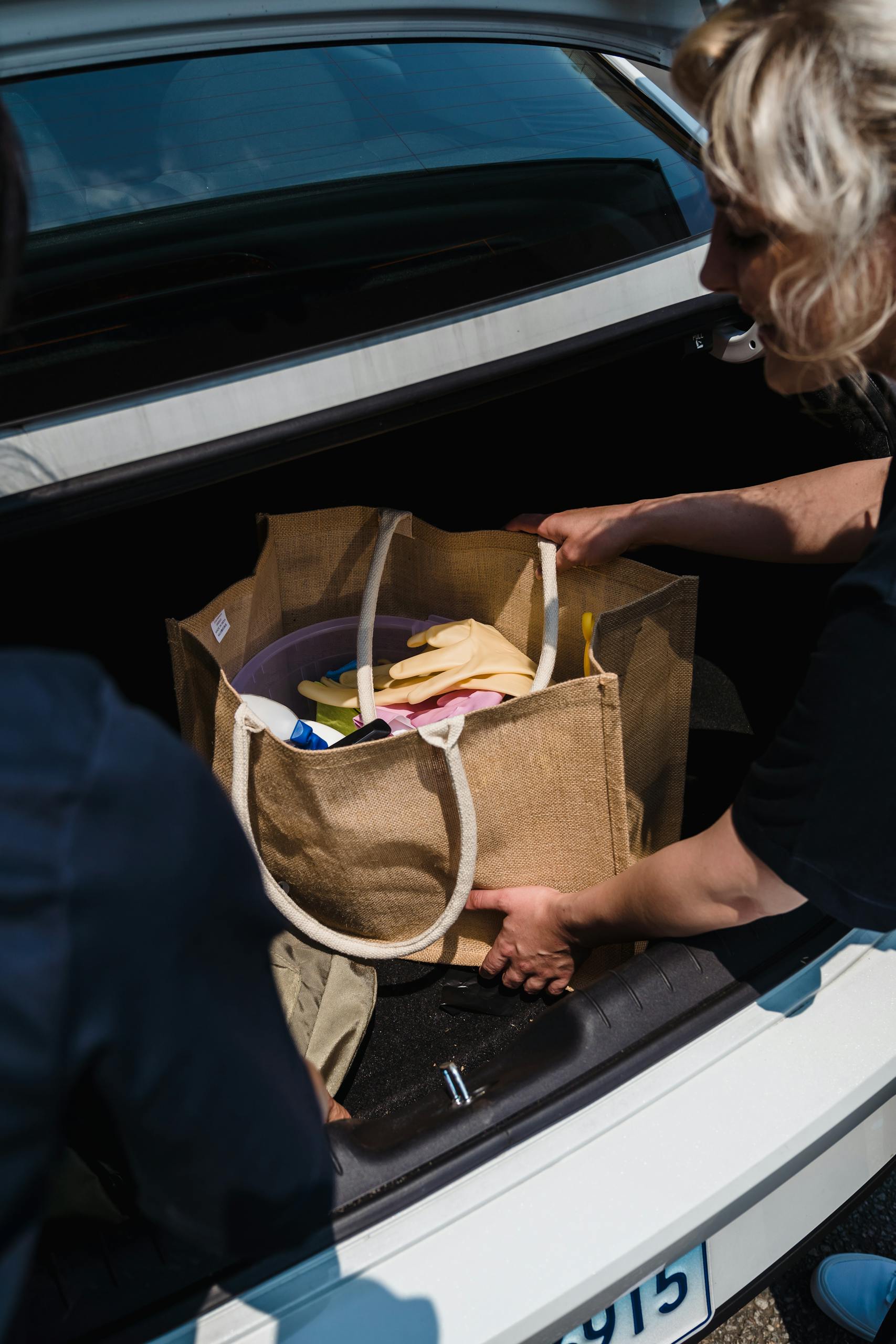 Two women pack cleaning supplies into a car trunk for transport.